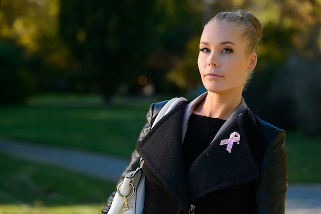 Portrait of young beautiful blond woman with pink ribbon for symbol of breast cancer awareness at the park