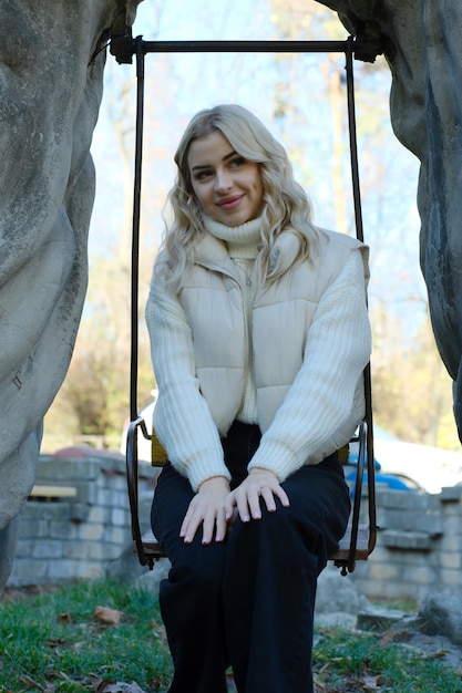 Portrait of a young beautiful blonde with long hair in a white sweater and jacket on a sunny autumn on a children's swing