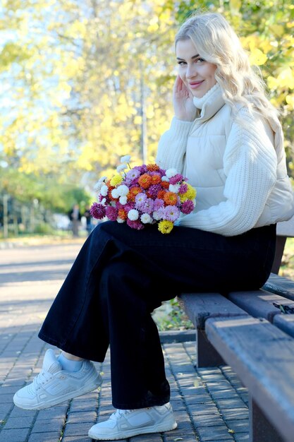 Portrait of a young beautiful blonde with long hair in a white sweater and jacket in sunny autumn in a city park with a bouquet of flowers