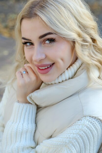 Portrait of a young beautiful blonde with long hair in a white sweater and jacket on a sunny autumn walk in the city