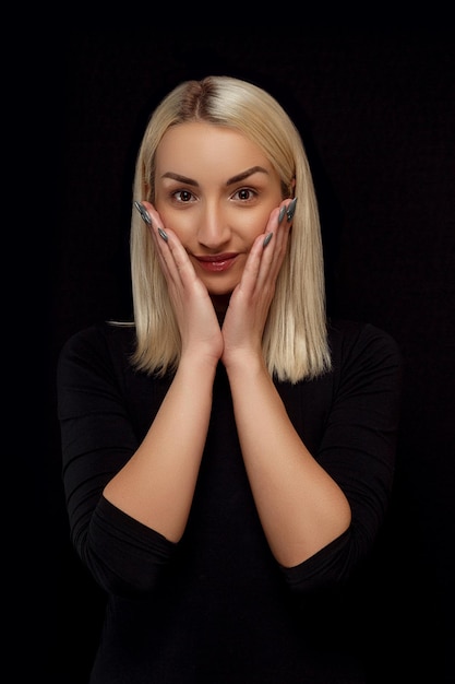 Foto ritratto di una giovane e bella donna bionda con le mani sul viso in abiti neri su sfondo nero isolato