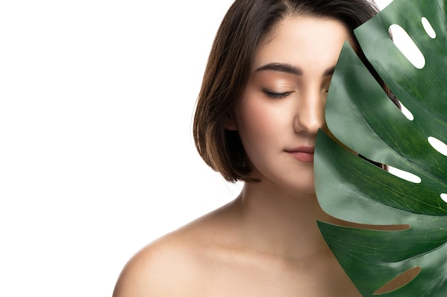 Portrait of young beautiful brunette woman with a monstera deliciosa tropical leaf on white background