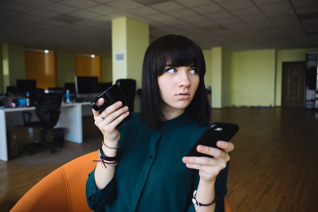 Portrait of a young, beautiful business woman who uses a mobile phone and drinking coffee. Break work. Modern office