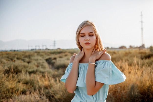 Portrait of young beautiful caucasian blonde girl in light blue dress standing on the field with sun-dried grass during sunset