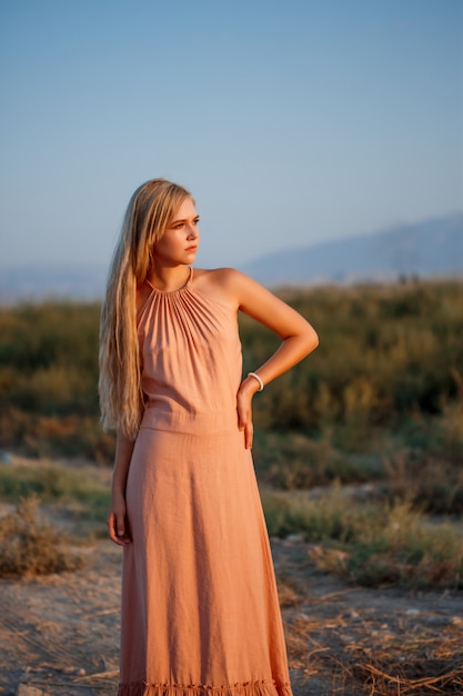 Portrait of a young beautiful caucasian blonde woman in a pink dress in a deserted field against the sunset