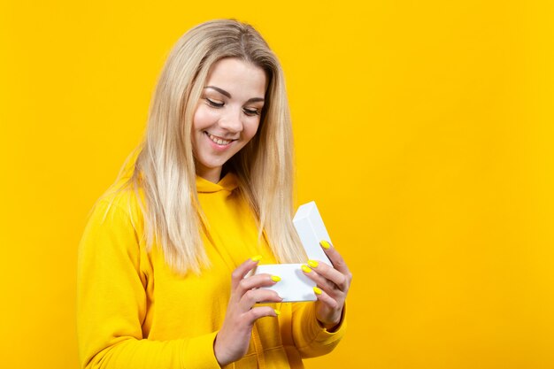 Ritratto di giovane bella donna bionda caucasica in abito sportivo giallo, con in mano una confezione regalo bianca,