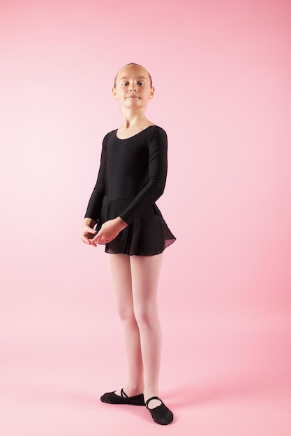 Portrait of young beautiful child girl ballerina standing practicing ballet wearing black tutu dress posing in studio with light pink background.