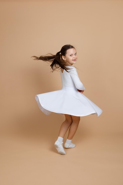 Portrait of young beautiful cute smiling girl with long dark hair in white dress socks and gymnastics shoes whirling in dance on brown background