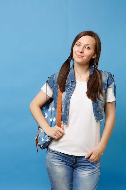 Portrait of young beautiful cute woman student in white t-shirt and denim clothes with backpack keeping hand in pocket isolated on blue background. Education in college. Copy space for advertisement.