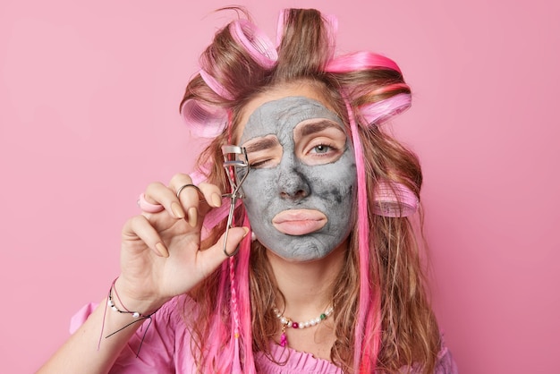 Portrait of young beautiful European woman applies clay mask uses eyelashes curler does makeup and beauty procedures isolated over pink background Women facial treatments and hairstyling concept