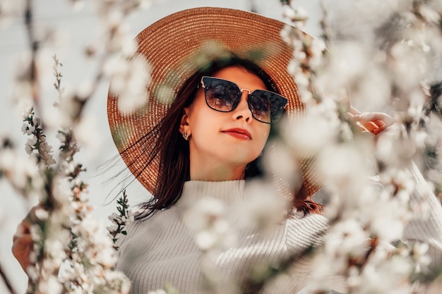 Portrait of young beautiful fashionable girl in hat posing near blooming tree with white flowers on a sunny day. Spring, girl near. 