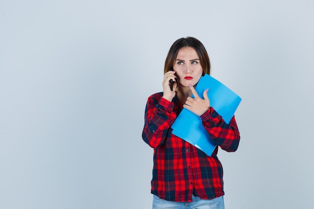 Portrait of young beautiful female holding folder while talking on phone, touching chin with finger in casual shirt, jeans and looking pensive front view