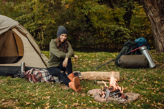 Portrait of young beautiful female tourist sitting on the log in the forest near tent and sleeping bag