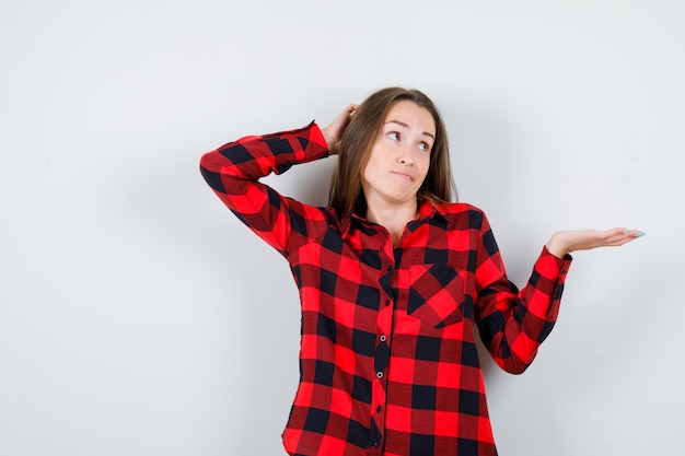 Portrait of young beautiful female with hand behind head, spreading palm aside in casual shirt and looking pensive front view