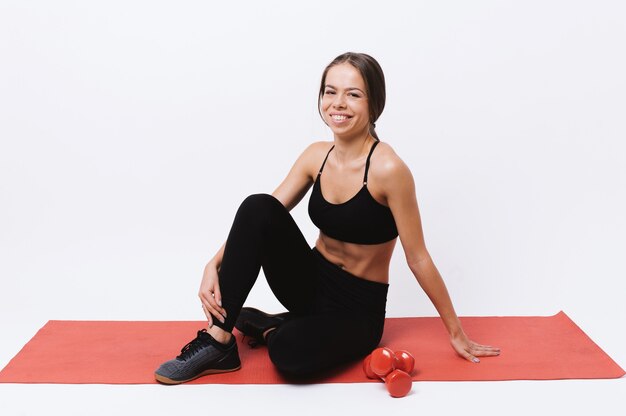 Portrait of young beautiful fitness woman sitting on red yoga mat and looking at the camera