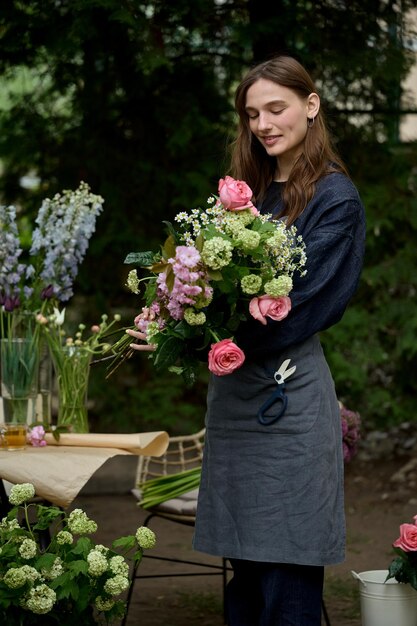 Portrait of young beautiful florist in apron on terrace making bouquet on green background