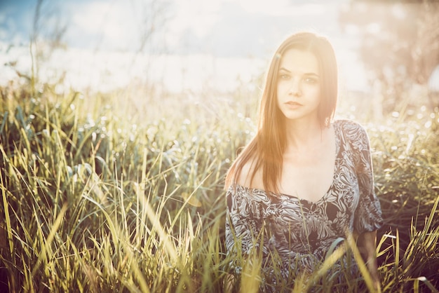 Ritratto di giovane bella ragazza in un vestito che si siede nell'erba durante il tramonto