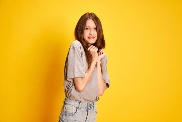 Portrait of young beautiful girl emotionally posing with fearful unpleasant face against yellow