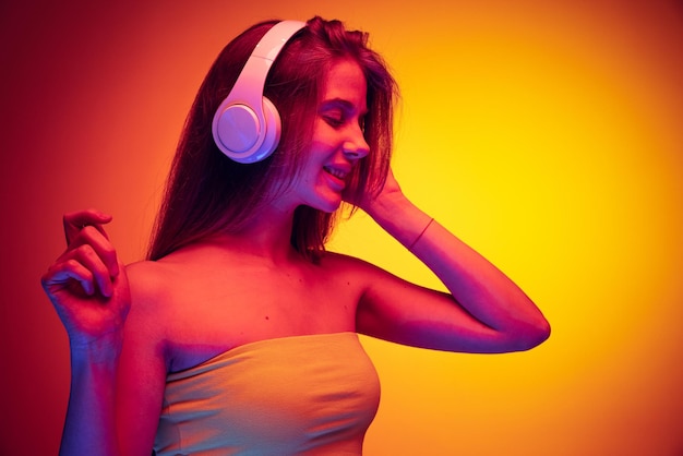 Portrait of young beautiful girl listening to music in headphones dancing isolated over gradient red yellow background in neon