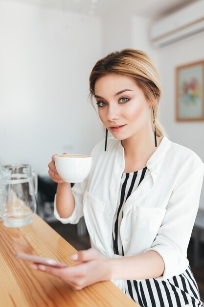 Portrait of young beautiful girl looking in camera with cup of coffee and mobile phone in hands at cafe. Pretty lady with blond hair sitting at the counter and drinking coffee in coffee shop