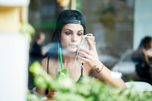 Portrait of a young beautiful girl. She sits alone in a cafe, listens to music on headphones and checks the make-up through the camera of a smartphone. Shot through the window.