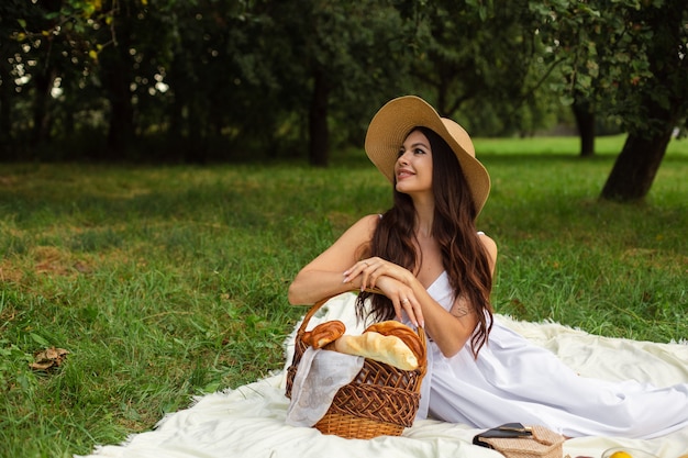 Портрет молодой красивой девушки с ровными белыми зубами, красивой улыбкой в соломенной шляпе и длинном белом платье на пикнике в саду