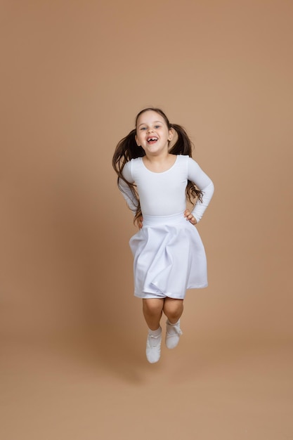 Portrait of young beautiful girl with long dark hair in white dress socks and gymnastics shoes jumping holding hands on waist on brown background