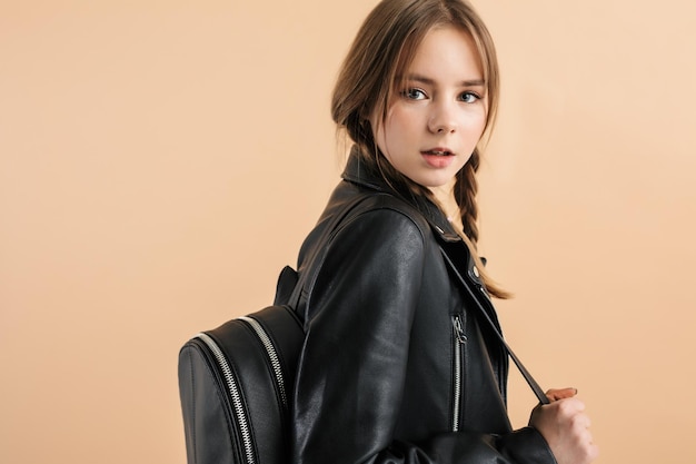Portrait of young beautiful girl with two braids in leather jacket with black backpack on shoulder while dreamily looking in camera over beige background