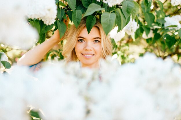 Photo portrait of young beautiful happy cheerful smiling positive blonde girl looking through branches with white flowers in summer blooming park.