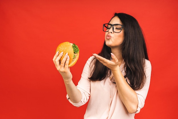 ハンバーガーを食べる若い美しい空腹の女性の肖像画 赤い背景の上にファーストフードを持つ学生の孤立した肖像画 ダイエットの概念