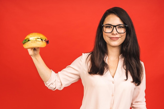 ハンバーガーを食べる若い美しい空腹の女性の肖像画 赤い背景の上にファーストフードを持つ学生の孤立した肖像画 ダイエットの概念