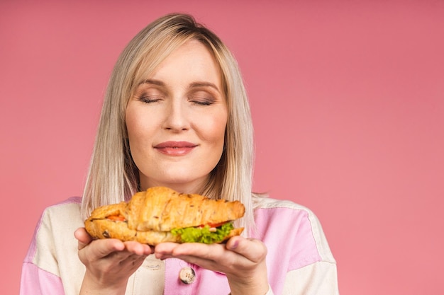 クロワッサンサンドイッチを食べる若い美しい空腹の女性の肖像画。ピンクの背景の上のファーストフードと女性の孤立した肖像画。ダイエットのコンセプト。