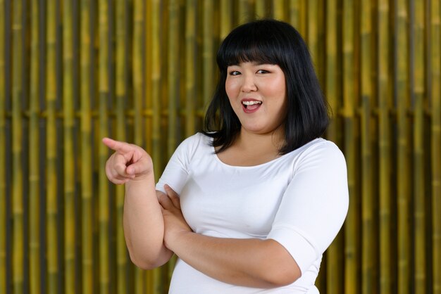 Portrait of young beautiful overweight Asian woman against bamboo wall outdoors