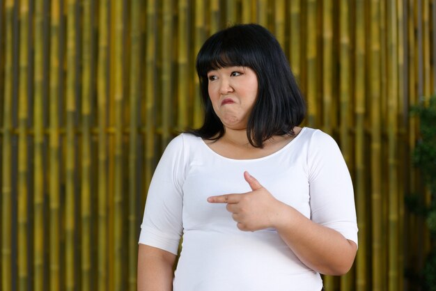 Photo portrait of young beautiful overweight asian woman against bamboo wall outdoors
