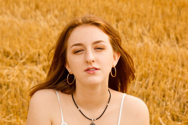 Portrait of a young beautiful redhead girl in the middle of a wheat field. He looks into the frame, brown eyes. Windy plays with hair. White cotton dress, eco style.