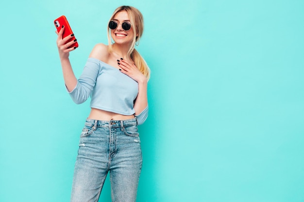 Portrait of young beautiful smiling blond female in trendy summer clothes sexy carefree woman posing near blue wall in studio positive model having fun indoors cheerful and happy taking selfie