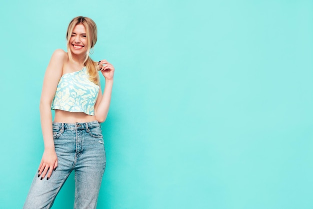 Portrait of young beautiful smiling blond female in trendy summer clothes Sexy carefree woman posing near blue wall in studio Positive model having fun indoors Cheerful and happy