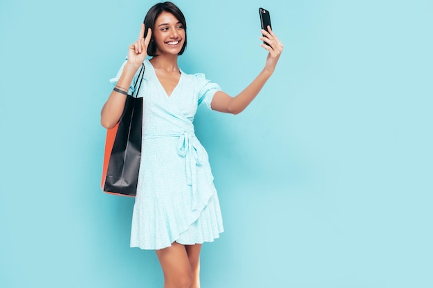 Portrait of young beautiful smiling female in trendy summer dress Carefree woman posing near blue wall in studio Positive model holding shopping bag Cheerful and happy Taking selfie