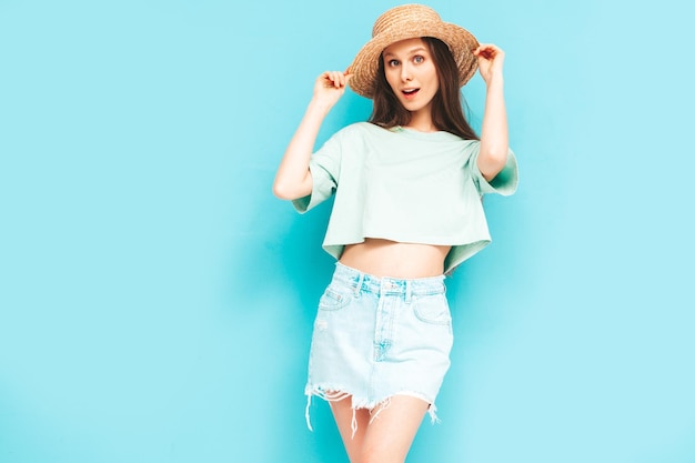 Portrait of young beautiful smiling female in trendy summer\
jeans skirt carefree woman posing near blue wall in studio positive\
model having fun indoors cheerful and happy in hat