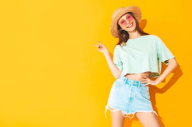 Portrait of young beautiful smiling female in trendy summer jeans skirt carefree woman posing near yellow wall in studio Positive model having fun indoors in sunglasses Cheerful and happy In hat