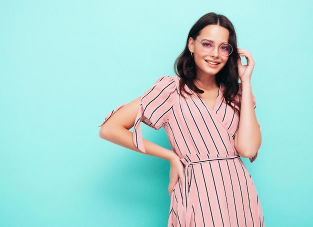 Portrait of young beautiful smiling female in trendy summer pink dress Sexy carefree woman posing near blue wall in studio Positive model having fun indoors Cheerful and happy in sunglasses