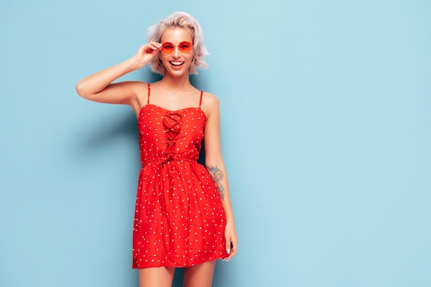 Portrait of young beautiful smiling female in trendy summer red dress Sexy carefree woman posing near blue wall in studio Positive blond model having fun and going crazy Cheerful and happy