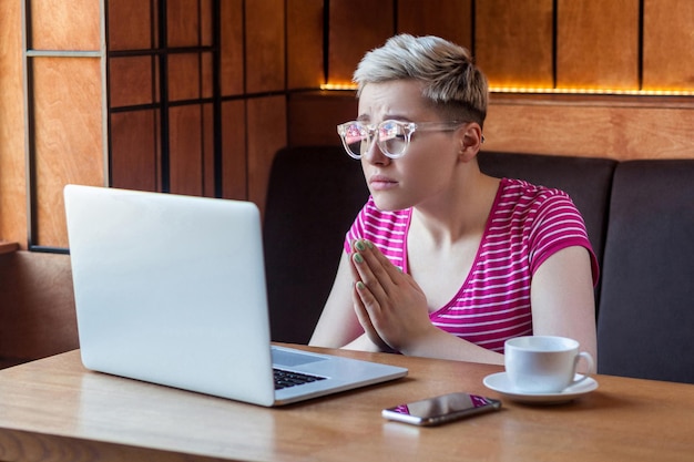 Portrait of young beautiful student with blonde short hair in pinlk tshirt is sitting in cafe and talking thought webcame with group mate begging for help with her homework indoor