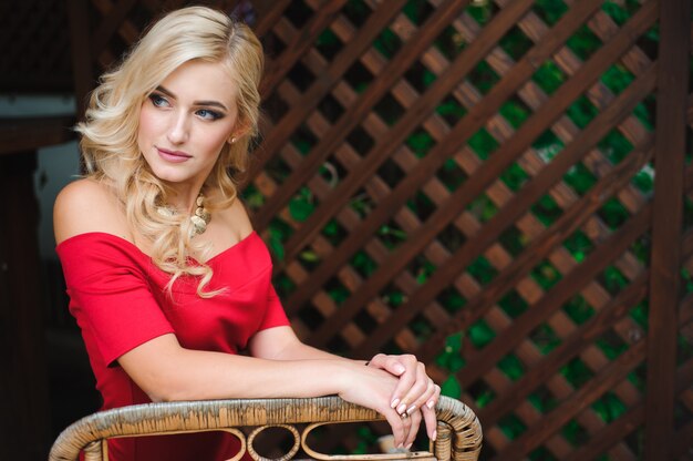 Portrait of young beautiful tanned blonde woman in red evening dress sitting outdoor in street cafe alone