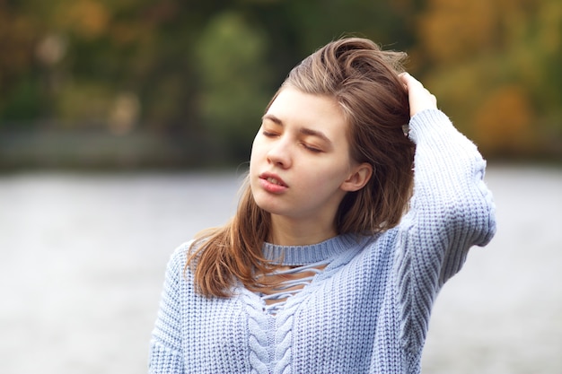 Portrait of young beautiful woman at autumn park with lake standing with eyes closed, enjoying good