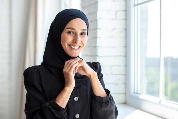 Portrait of a young beautiful woman in a black burqa hijab standing by the window smiling at the