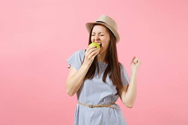 Portrait of young beautiful woman in blue dress, summer straw hat holding, eating green fresh apple fruit isolated on pink background. Healthy lifestyle, people, sincere emotions concept. Copy space.