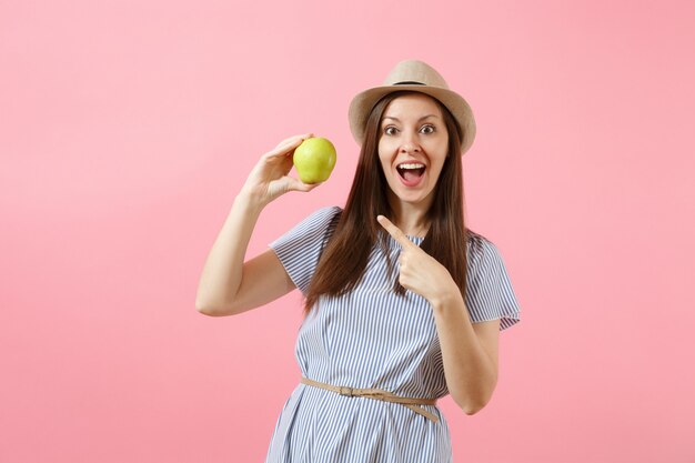 Portrait of young beautiful woman in blue dress, summer straw hat holding, eating green fresh apple fruit isolated on pink background. Healthy lifestyle, people, sincere emotions concept. Copy space.