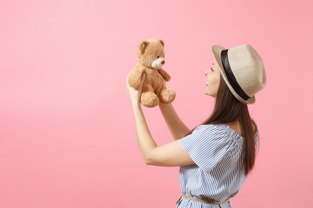 Portrait of young beautiful woman in blue dress, summer straw hat holding teddy bear plush toy isolated on pink background. People, sincere emotions, lifestyle concept. Advertising area. Copy space.