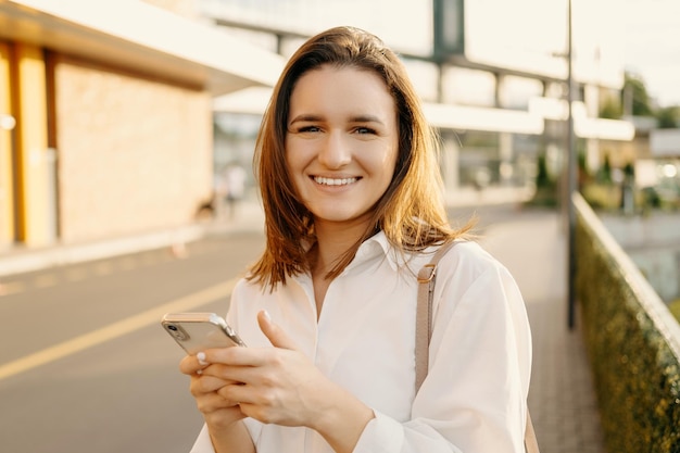 カメラを見て、スマート フォンの街の背景を保持しているカジュアルな屋外で若い美しい女性の肖像画